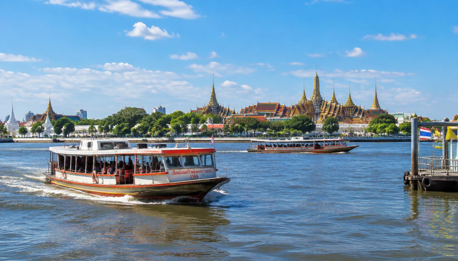 Ferry boats on the Chao Phraya River in daytime, with the golden spires of Bangkok's Grand Palace on the riverbank
