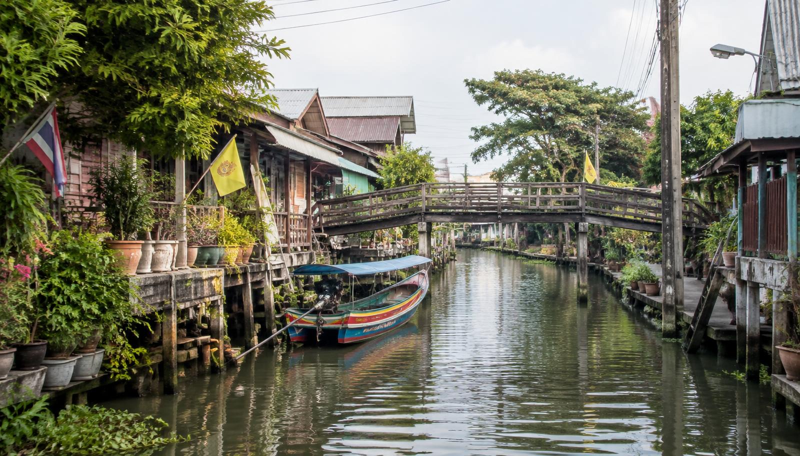 Quiet canal in a Bangkok neighbourhood with wooden stilt houses, potted plants, a wooden footbridge and a moored long-tail boat